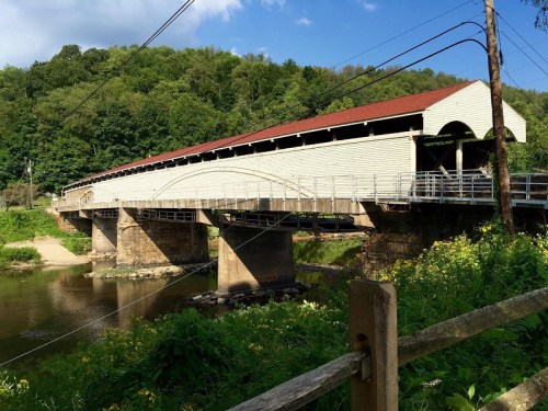 A covered bridge spans a river, surrounded by lush greenery and a clear blue sky.