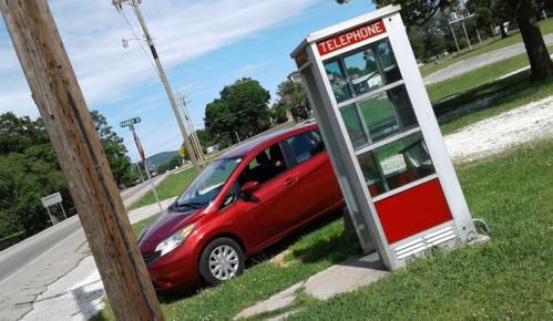 A red car parked near an old telephone booth on a grassy roadside. Trees and a power pole are in the background.