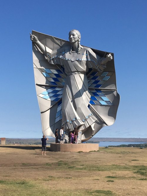 A large metallic statue of a woman in a flowing dress, with blue accents, stands on a grassy area with people nearby.