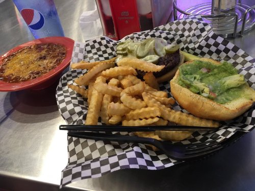 A plate with a burger topped with green sauce, crinkle fries, and a bowl of chili on a checkered paper.