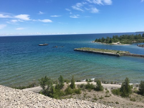 A serene view of a blue lake with green shores and a clear sky, featuring rocky areas and distant land.
