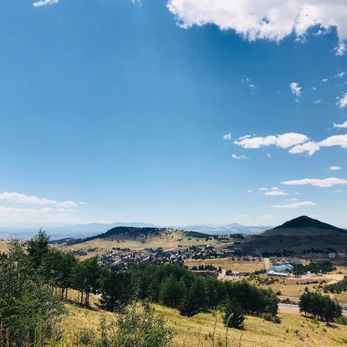 A scenic landscape featuring rolling hills, green trees, and a clear blue sky with scattered clouds.