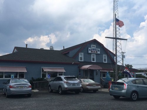 Exterior of Kubi's Bar, a blue building with a flagpole, surrounded by parked cars under a cloudy sky.