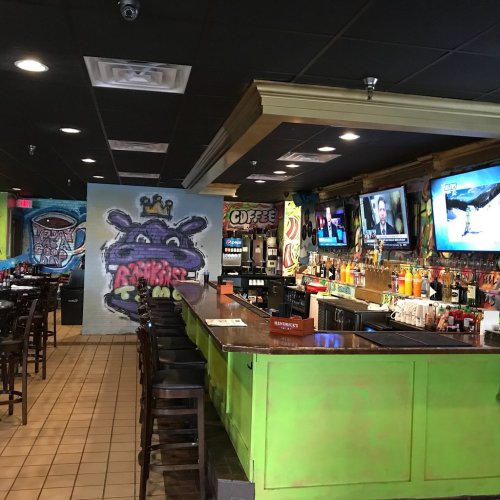 A vibrant bar area with colorful decor, TVs, and a mural, featuring seating and a variety of drinks on display.