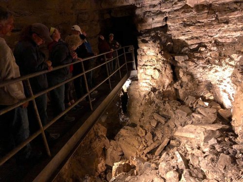 Visitors stand on a walkway in a cave, observing rocky walls and a partially collapsed area illuminated by soft lighting.