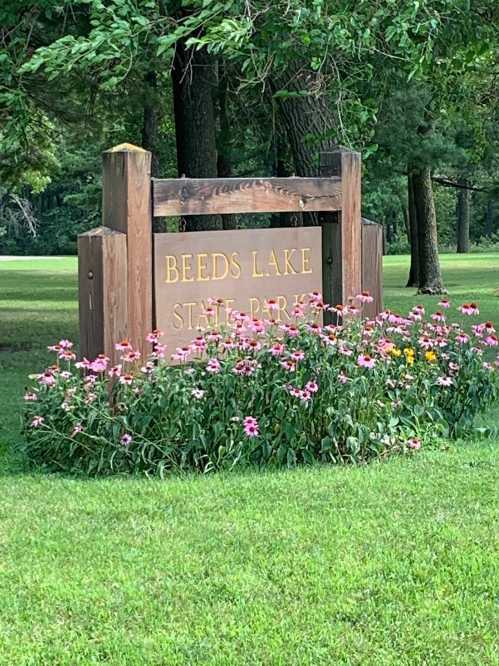 Sign for Beeds Lake State Park surrounded by colorful flowers and green grass in a wooded area.