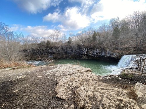 A serene landscape featuring a river and waterfall, surrounded by bare trees and a cloudy sky.
