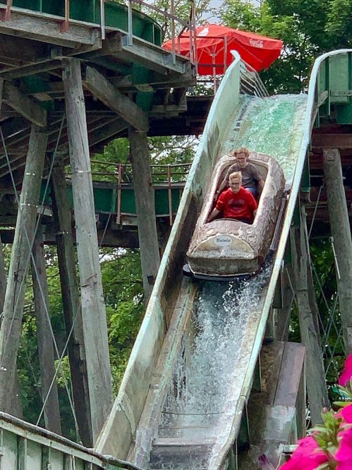 Two people enjoy a water ride, splashing down a wooden slide surrounded by greenery and colorful flowers.