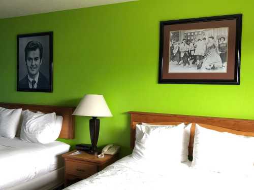 A brightly colored hotel room with two beds, a lamp, and framed black-and-white photos on the green wall.