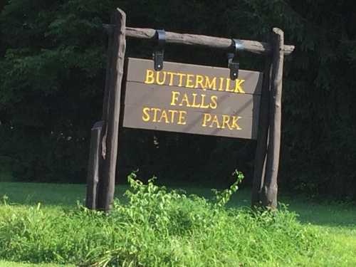 Sign for Buttermilk Falls State Park, featuring wooden posts and yellow lettering against a natural green backdrop.