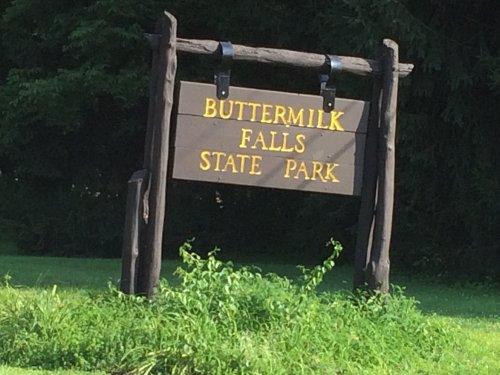 Sign for Buttermilk Falls State Park, featuring wooden posts and yellow lettering against a natural green backdrop.