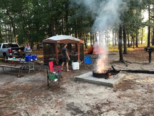 A campsite with a fire pit, tents, and people preparing food in a wooded area. Smoke rises from the fire.