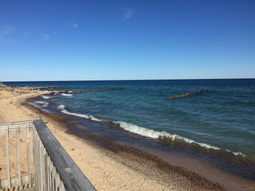 A serene beach scene with gentle waves, sandy shore, and a clear blue sky. Wooden posts are visible in the water.