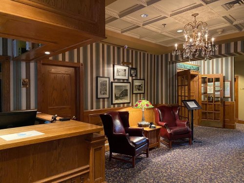 Cozy lobby with wooden accents, striped wallpaper, two armchairs, a chandelier, and framed photos on the walls.