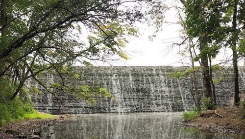 A serene waterfall cascading over a stone dam, surrounded by lush greenery and reflected in a calm pond.