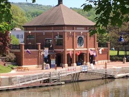 A brick building with a round window, flags, and a dock by a calm waterway, surrounded by greenery.