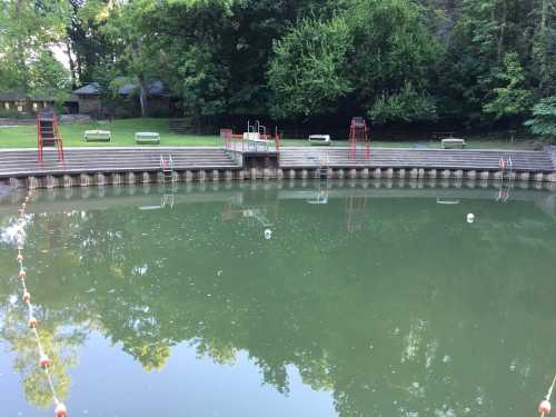 A calm pond surrounded by trees, with a wooden dock and red ladders leading into the water. Benches are visible nearby.