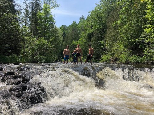 Four people stand on rocks in a river, surrounded by lush greenery and a waterfall, enjoying a sunny day outdoors.
