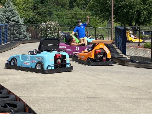Two go-karts on a track, one blue and one orange, with a person in a blue shirt waving. Trees in the background.