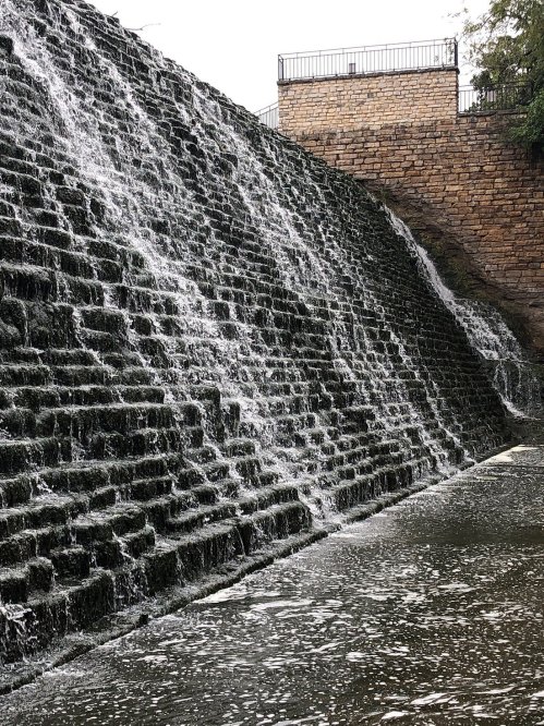 A cascading waterfall flows down a stepped stone structure into a calm body of water below.