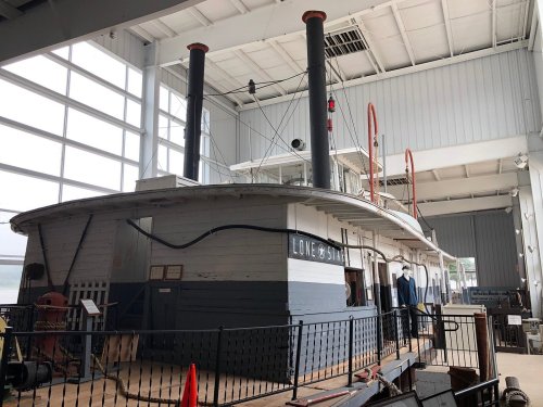 A historic steamboat displayed indoors, featuring two tall smokestacks and a wooden hull, with large windows in the background.