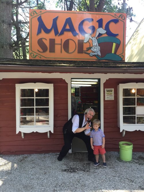 A magician poses playfully outside a colorful magic shop, with a child standing beside him.