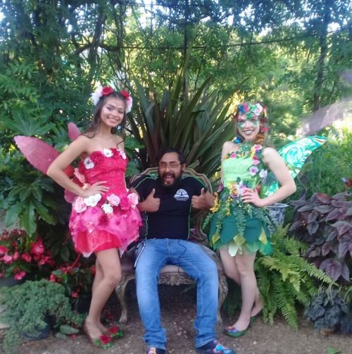 Two women in colorful fairy costumes with flowers pose beside a man sitting on a bench in a lush garden.
