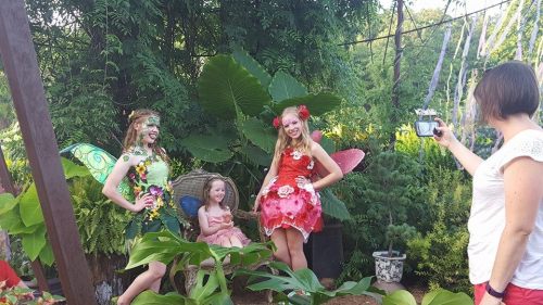 Two girls in fairy costumes pose with a third girl, while an adult takes a photo in a lush garden setting.