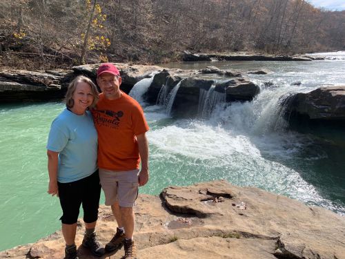 A couple stands on a rock by a turquoise river with a waterfall in the background, surrounded by trees.