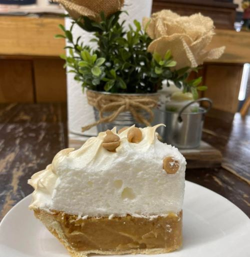 A slice of creamy dessert topped with meringue and nuts, served on a white plate with a floral arrangement in the background.