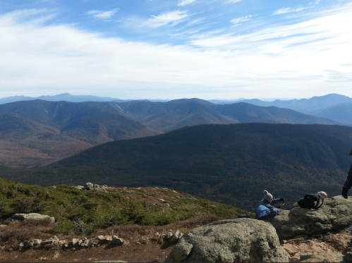 A panoramic view of mountains and valleys under a blue sky, with hikers resting on rocky terrain in the foreground.