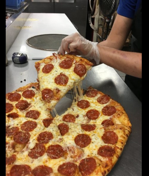 A hand in a glove lifts a slice of pepperoni pizza, with cheese stretching from the pie on a metal countertop.