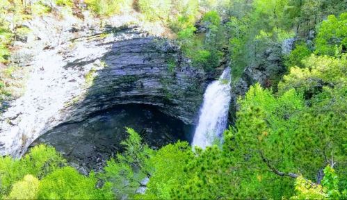 A waterfall cascades down rocky cliffs surrounded by lush green trees in a serene natural landscape.