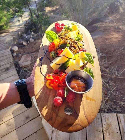 A wooden platter with colorful fruits, herbs, and a small bowl of sauce, held outdoors in a natural setting.