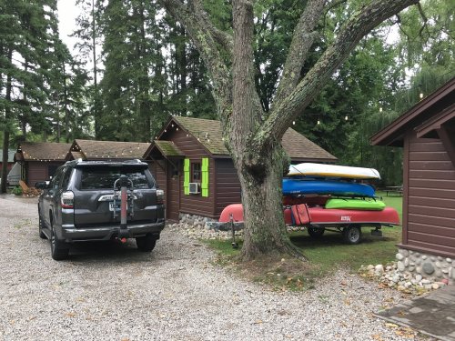 A gray SUV parked near brown cabins, with colorful kayaks stacked on a trailer under a tree.