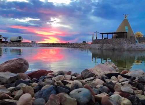 A serene landscape featuring a pond reflecting a colorful sunset, with a teepee and rocky shoreline in the foreground.