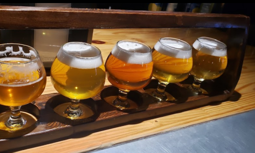 A wooden flight holder with five small glasses of different beers, showcasing various colors and foam levels.
