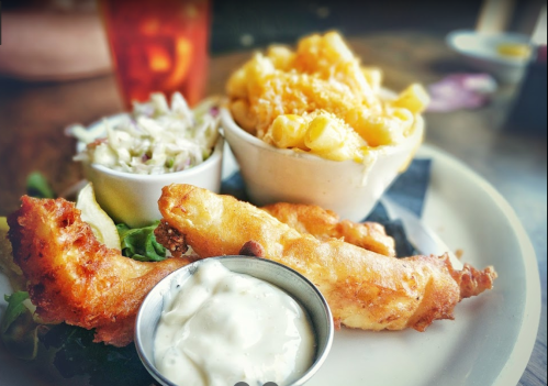 A plate with two crispy fish fillets, creamy coleslaw, macaroni and cheese, and a small bowl of dipping sauce.