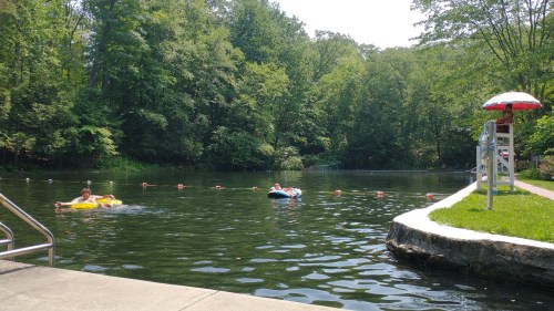 Two people float on inner tubes in a calm, green lake surrounded by trees on a sunny day. A lifeguard stand is nearby.
