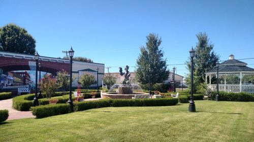 A sunny park featuring a fountain, greenery, and decorative lampposts, with a gazebo in the background.