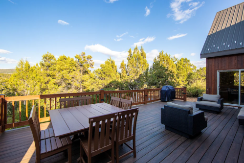 A wooden deck with a dining table and chairs, overlooking trees and a clear blue sky.