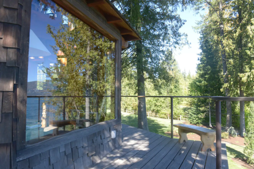 A wooden deck with a railing overlooks a lush green landscape and trees, reflecting sunlight through large windows.