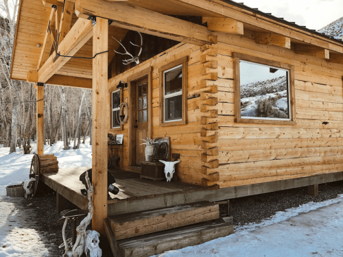 A rustic wooden cabin with a porch, surrounded by snow and featuring decorative antlers and a wagon wheel.