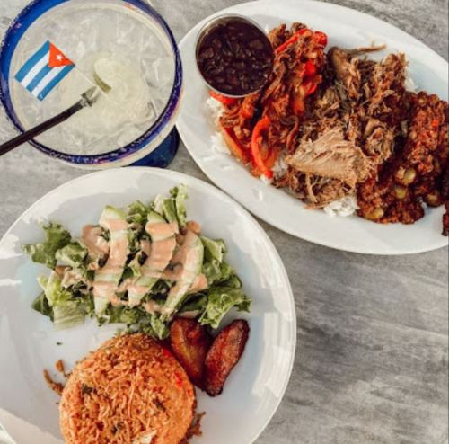 A plate of Cuban food featuring rice, salad, plantains, and shredded meat, alongside a cocktail and a Cuban flag.