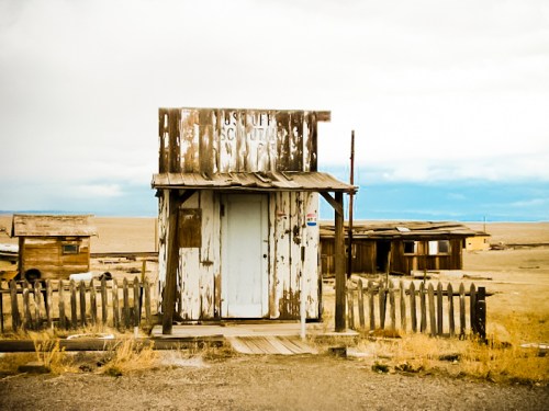 A weathered wooden shack with a faded sign, surrounded by a barren landscape and distant buildings under a cloudy sky.