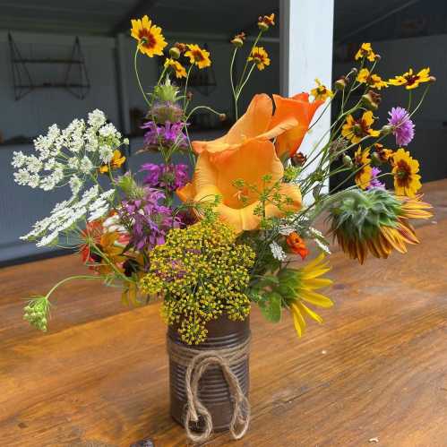 A vibrant bouquet of wildflowers in a rustic can, featuring orange roses, sunflowers, and various colorful blooms.