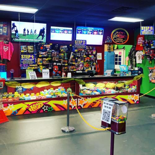 Brightly colored arcade with gaming stations, merchandise displays, and screens showing gameplay. Queue area with ropes.