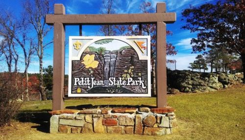 Sign for Petit Jean State Park featuring a waterfall illustration and autumn leaves, set against a natural landscape.