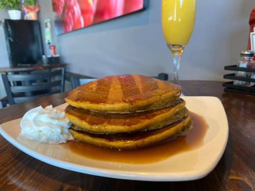 A stack of three golden pancakes drizzled with syrup, served with whipped cream and a mimosa in the background.