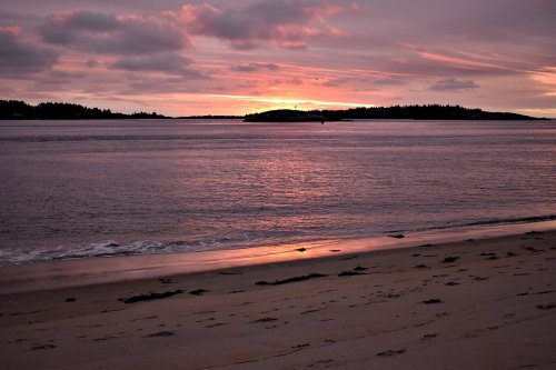 A serene beach at sunset, with soft pink and purple hues reflecting on calm water and distant islands.